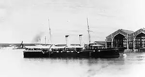 A large warship with a black hull moored in harbor, with canvas awnings covering the deck