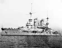 A large warship at rest, with light gray smoke drifting up from its two smokestacks