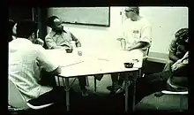 B/w photo of several white and one black person who are sitting at a table with a whiteboard on the wall behind. They are all looking to one white male person sitting at the table wearing a white t-shirt with a number on the chest.