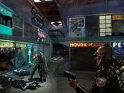 A green-cloaked man is staring from the right corner through black goggles to another deformed human being, who is close to a dead body of a woman laying on the floor along with several blood marks and some destroyed things.