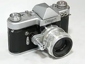 A photograph of the silver-black camera's front, on a white table, lighted from above