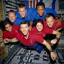 Seven crew members, in red or blue collared shirts, floating in microgravity.