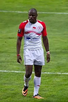 A man wearing a rugby uniform consisting of a white shirt with red sleeves, white shorts, and white socks catches his breath standing on a rugby pitch.