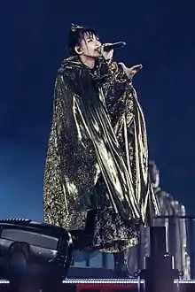 A girl draped in a black and gold robe with a microphone in her left hand, and her right hand outstretched