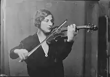 A young white woman playing a violin indoors. She has hair in marcelled waves, and is wearing a dark garment with a deep V neckline