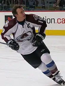 A hockey player with short, brown hair looks to his left as he skates. He is in a white and burgundy uniform with a stylized A logo on his chest.