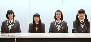 Four girls wearing school uniform outfits sit at a table against a white background