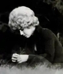 A young woman with blonde curly hair, wearing dark clothing and sitting on grass