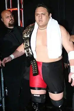 An adult male wearing red and black tights with black wrestling boots holding a black championship belt.