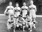 A group of seven young men arranged in two rows—four standing in back and three squatting in front—in basketball uniforms: white tank tops with a horizontal stripe and interlocking "SC" logo and white colored shorts.