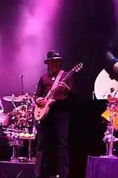 A man wearing a hat and dark clothing playing a guitar on stage