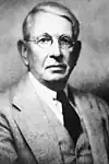 A photographic portrait of a man wearing glasses and a suit