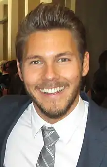 A man with dark hair wearing a grey suit including a red and grey pattern tie.