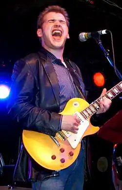 Sean Costello wearing a blue striped shirt, black leather jacket, and blue jeans, standing onstage, playing an electric guitar and singing into a microphone