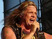 A middle-aged Caucasian man with long, blond hair and tattoos on his right shoulder wearing a sleeveless khaki shirt sings into a microphone.