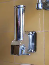 A vertical stainless steel tube, mounted on a wall, with a crank handle on the side at the bottom, next to the hopper-like horizontal opening from which the grated soap will fall