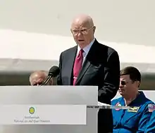 A bespectacled Glenn speaking at an outdoor podium