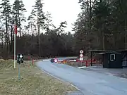 Guardpost with vehicle access control barrier, and red 'danger' flag aloft its flagstaff.  The circular signs on the pole to the right indicate 'no pedestrians' (top) and 'no vehicles' (bottom).