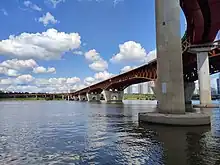 View from a bike trail below the new Seongsu bridge. The bridge is red and supported by concrete pillars. The sky is mostly clear with a few clouds and bright.