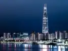 A nighttime shot of the Lotte tower from across the river. The tower is lit up and is far taller than any of the apartment buildings around it. By the riverside, a row of streetlights are shining on the river.