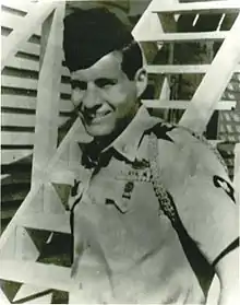 Head and torso of a smiling young man standing in front of a wooden staircase, wearing a short-sleeved military shirt with shoulder cords and a garrison cap.