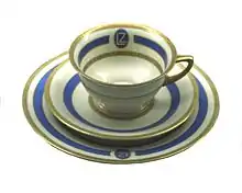 An elegant drinking cup on a saucer. Both are in white ceramic with pale blue and gold decoration.