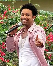 A man holding a microphone with one hard and extending his index finger with the other; he is wearing a white undershirt, red- and white-striped dress shirt and jewelry accessories. In the background is a man and vegetation.