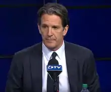 Brendan Shanahan stands behind a podium that has a LeafsTV microphone attached on its top.