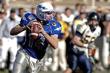 A uniformed American football player with the football looks ahead.