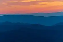 Five mountain ridges in shades of dark blue below an orange and yellow sunset.