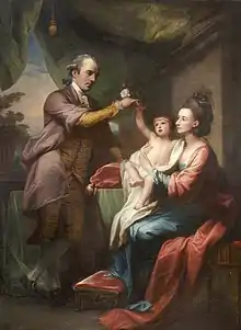 seated woman holding cherubic baby; man standing beside them placing flower in baby's hand