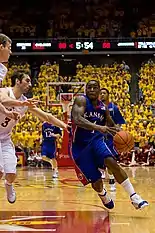 A person wearing a blue-colored basketball uniform dribbling a basketball running against another person wearing a white basketball uniform.