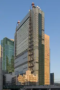 Ground-level view of a blue, glass, rectangular high-rise; attached to one side of the building are two structures consisting of poles that run the height of the building