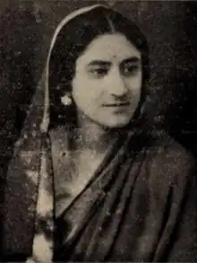 A South Asian woman with fair skin and dark hair parted center, wearing a scarf draped loosely over her head