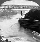 A woman is crossing on a rope over a river.  She wears a wide-brim hat and holds a pole to balance herself while her feet are in buckets. A double-deck bridge, filled with an audience on the lower deck, is in the background.