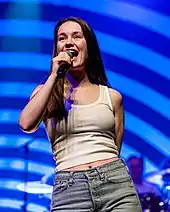 A woman wearing a white tanktop and blue jeans. She is singing into a microphone.