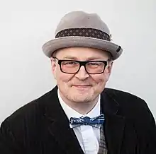 middle aged white male wearing hat and glasses, with shirt, vest, jacket, and bowtie, smiling at camera