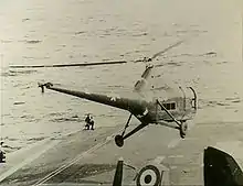 The forward section of an aircraft carrier's flight deck. A helicopter with United States markings and the letters "UP-28" painted on the side is hovering just above.