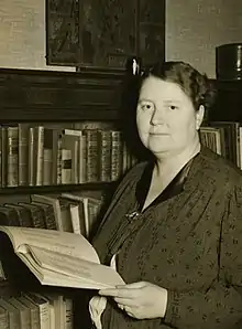A middle-aged white woman in a dark print dress, standing in front of a bookcase, holding a book.