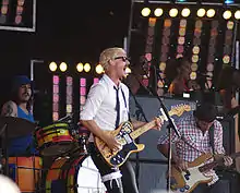 Gillies at left behind his drum kit. Johns is singing wide-mouthed into a microphone and playing his guitar, he wears a garter with a ribbon over his pants on his right thigh. Joannou is playing his bass guitar. In the background are three female singers with one partly obscured at right.