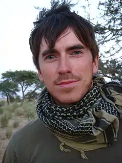 Portrait photograph of Simon Reeve; a white male. A BBC publicity photo, taken on location during his travels for the 'Tropic of Cancer with Simon Reeve' television series. Showing his head to his chest to just below shoulder level, Reeve, looking straight at the camera, has an engaging but neutral look on his face. Mouth closed, eyes blue-green topped with thick dark brown eyebrows, head covered in slightly unkempt dark brown hair, his face and top lip covered in very short ginger coloured stubble. He wears a green T-shirt, with an Arabic style head scarf loosely wrapped around his neck.