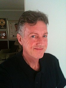 A fifty-five year-old man, shown in upper body shot. He is posed in front of a book shelf. He wears a dark top and is turned slightly towards the viewer.