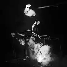 A black and white photo on the right side profile of the artist performing at a keyboard and surrounded by instruments. Smoke or fog is present across the floor and above.