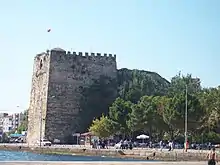 Photograph of a tall, roughly square stone fortress in a modern coastal city.