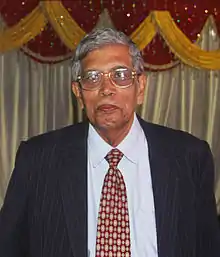 A photograph of an old man wearing blue suit and glasses.