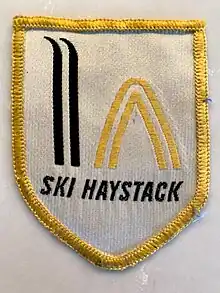 White arm patch showing a stylized pair of black skis held vertically next to a stylized yellow haystack, with the words "Ski Haystack" underneath