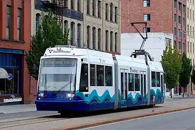 A short train traveling in the center of a street, passing several multi-story buildings.