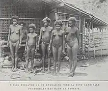 Black-and-white photograph of five nearly naked young women and girls standing in front of a hut; a sixth is visible in the background