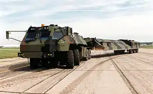 Long vehicle in green black and dirt brown camouflage executing a turn on light-colored paved road for camera.