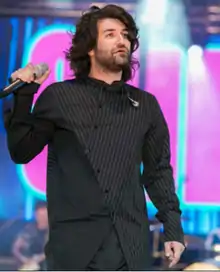 A man is seen wearing black clothing and holding a microphone in his hand as he stands in front of a blue-purple backdrop.